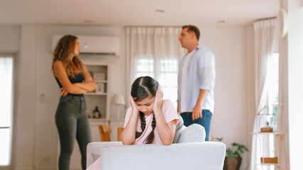 Annoyed and unhappy young girl sitting on sofa trapped in middle of tension by her parent argument...