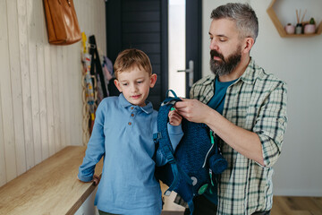 Father helping son get ready for kindergarten, preschool. Putting backpack on his back.