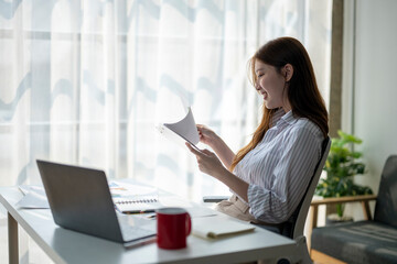 A woman is sitting at a desk with a laptop and a red mug. She is reading a piece of paper and smiling. Concept of productivity and relaxation