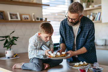 Boy filling pancake with fruit, sweets. Father spending time with son at home, making snack...
