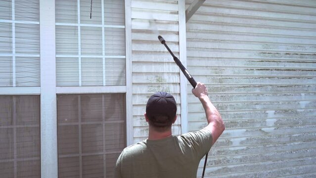 Homeowner pressure washes vinyl siding on hot day. Behind