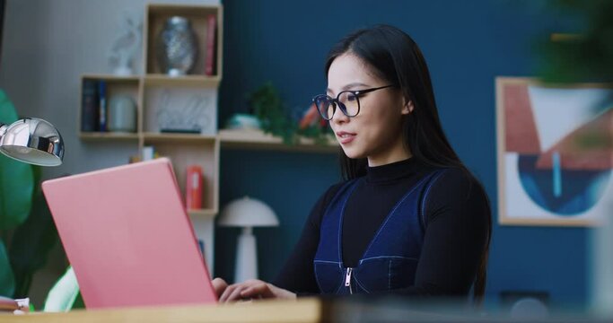 Beautiful Chinese girl sitting in front of her laptop. Woman giggling and laughing while writing something funny or replying to friend. Typing text on keyboard. Wearing glasses for better vision.