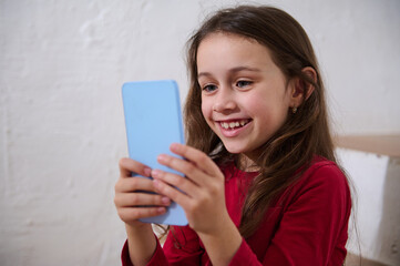 Naklejka premium Close-up portrait of a smiling happy child having fun on smart mobile phone, using mobile apps, playing online video games or watching movies webinars or kids bloggers, isolated white background
