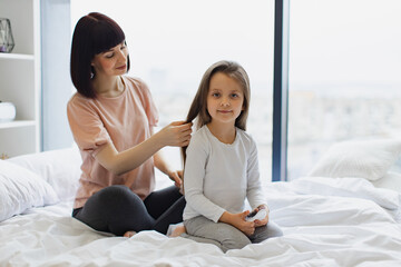 Charming little girl smiling while her cheerful mom brushing daughter's hair. Beautiful young...