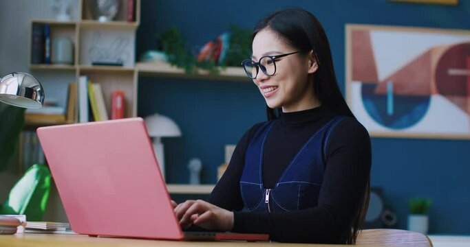 Side view of beautiful Asian girl sitting in front of laptop while entering text on keyboard. Smiling to herself from funny replies or ideas. Working remotely. Chatting with friend or relative.