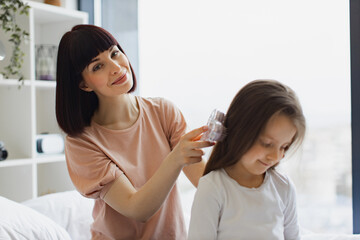 Charming Caucasian mother combing her daughter's hair while sitting on bed near big panoramic window. Happy family spending weekend day home.