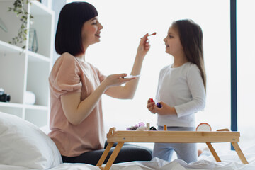Happy woman in casual clothes applies makeup with brush on the face of little girl while sitting in bed near panoramic window. Family, mother and daughter relax using cosmetics on weekend.