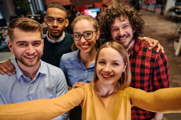 A diverse group of business professionals take a break from their tasks in a modern startup office to capture a creative selfie, showcasing teamwork and a vibrant workplace culture