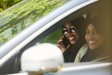 Two Muslim women wearing hijab converse on a smartphone while traveling together in a car through the 