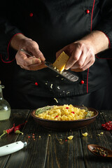 A professional chef grates hard cheese onto a plate with pasta using a hand grater. Working environment in a hotel kitchen with spices and kitchen utensils.