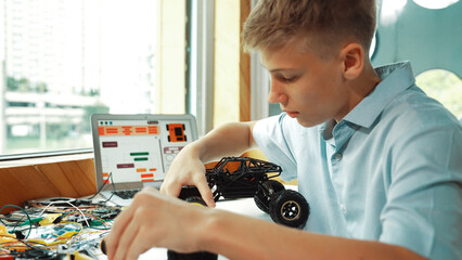 Happy teenager fixing robotic model while using electronic equipment remove car wheel with wire and laptop scatter around. Smart boy study or learn about car robot construction at class. Edification.