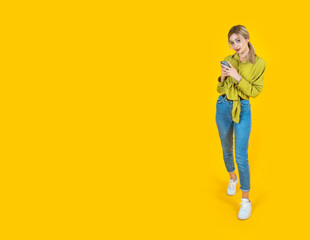 Fototapeta premium Full body size blonde 20s caucasian woman hold use mobile phone device. Looking camera. Yellow background, copy space. Online shopping using app on smartphone. Social media design concept image.