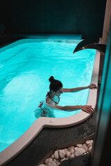 A young woman in a swimsuit poses in an illuminated indoor pool in a hotel. Hotel holiday concept....