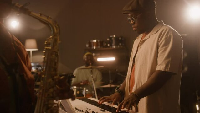 Medium slowmo with lens flare of unrecognizable African American woman playing saxophone, black men playing keyboards and drums, while performing together in studio or at club gig