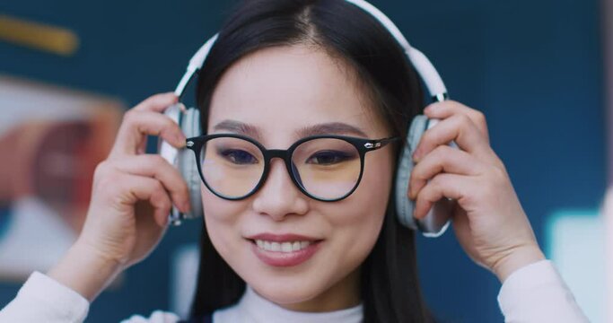 Facial view of Asian girl. Woman looking very focused at something. Putting on headphones. Enjoying music while looking at screen or reading book. Wearing large glasses for better vision.