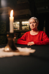 Vertical portrait of grey-haired adult woman receiving forecasting future during divination session sitting at table in dark room, by light of burning candle, looking at camera with serious expression