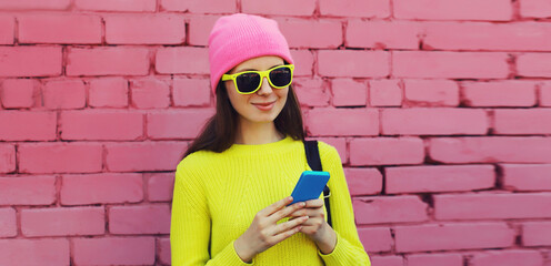 Modern young woman with mobile phone looking at device wearing colorful vivid clothes on city street