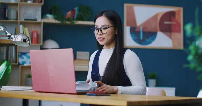Cute Chinese woman sitting in front of rose laptop. Thinking what to do. Deciding to listen to music. Putting on big grey headphones. Enjoying favourite song. Relaxing weekend. Taking break from work.
