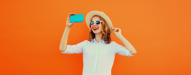 Portrait of happy smiling young woman taking selfie with smartphone wearing straw hat on orange
