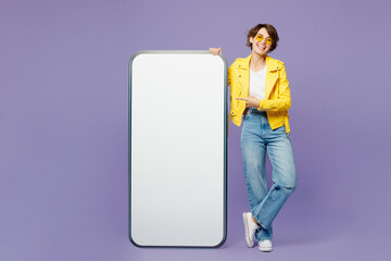 Full body young fun woman she wears yellow shirt white t-shirt casual clothes glasses point finger on big huge blank screen mobile cell phone smartphone with area isolated on plain purple background.