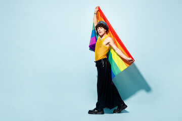 Full body side view young happy gay Latin man wear mesh tank top hat clothes hold rainbow flag isolated on plain pastel light blue background studio portrait. Pride day June month love LGBT concept.