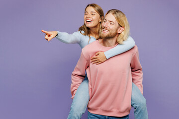 Young couple two friends family man woman wear pink blue casual clothes together giving piggyback ride to joyful sit on back point finger aside isolated on pastel plain light purple background studio