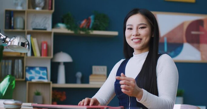 Cute Chinese female working or studying remotely. Pressing keys on keyboard of pink laptop. Shutting down computer. Closing deck lid with one hand. Looking directly at camera and showing heart symbol.