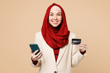 Young surprised Arabian Asian Muslim woman in red abaya hijab suit clothes hold mobile cell phone credit bank card shopping online isolated on plain beige background UAE middle eastern Islam concept
