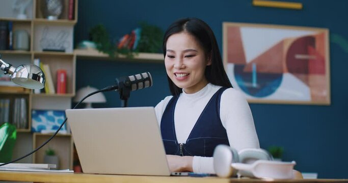 Cute Asian woman sitting at her desk. Talking to big microphone besides laptop. Looking at screen and waving to someone. Online tutor or student. Concept of working or studying remotely.
