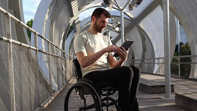 Middle-aged man in wheelchair engrossed in using a tablet, emphasizing accessibility in a modern urban setting. Focused Man in Wheelchair Using Tablet on Architectural Bridge