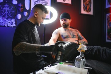 bearded tattoo artist working at his studio tattooing sleeve on the arm of his male client. Man getting tattooed by professional tattooist