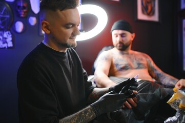 Male tattoo artist holding a tattoo gun, showing a process of making tattoos on a male tattooed model's arm