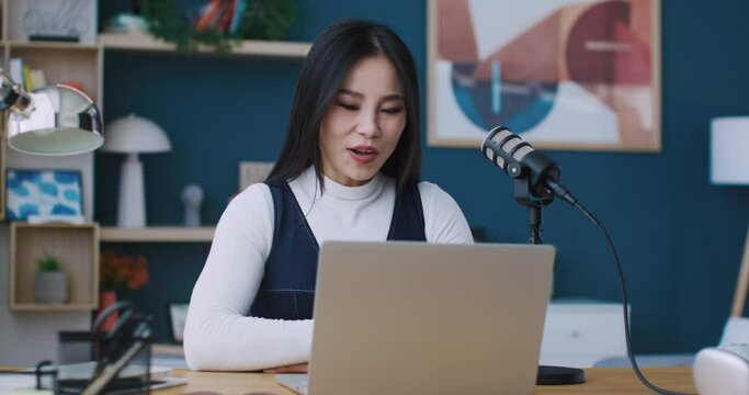 Beautiful Asian woman sitting at desk in front of laptop. Teacher waving to students. Teaching remotely. Using professional microphone setup for sound quality. Talking with video camera on.