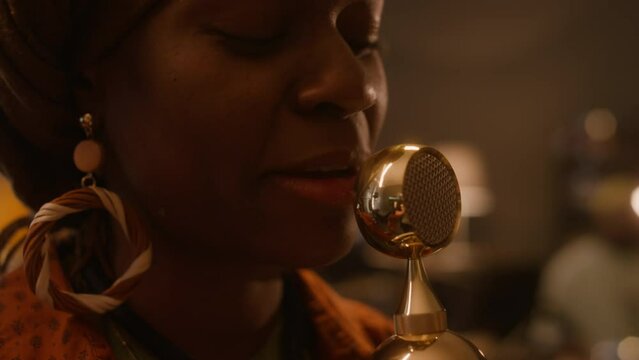 Extreme close-up of African American ethnic band singer in turban, with large earrings giving emotional performance, singing into golden vintage microphone, while playing gig with band in club