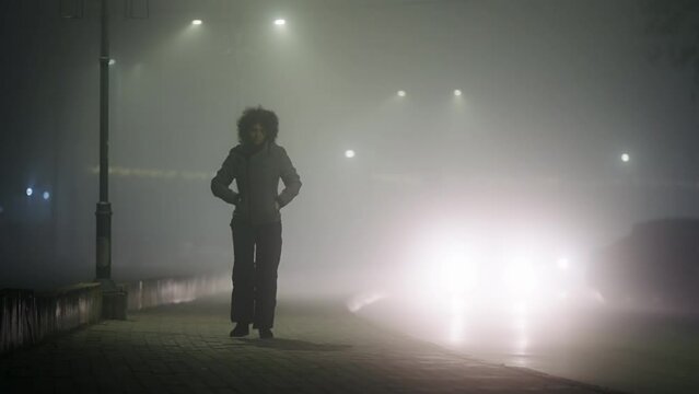 A woman is walking down a sidewalk pavement in the dark foggy time. The street is lit by street lamps. The woman is wearing a white jacket and she is alone. The scene is quiet and peaceful
