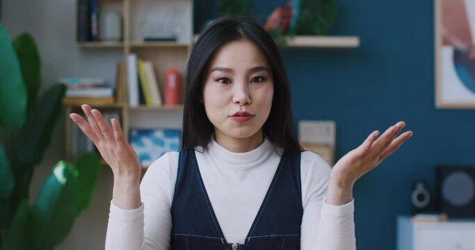 Short conversation with cute Asian female. Girl looking directly at camera. Beginning to wave and smile. Explaining some process by using hand gestures. At end showing heart symbol with two fingers.