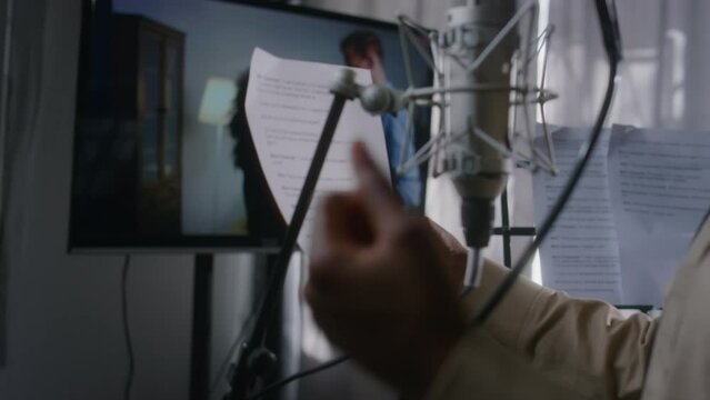 Medium close-up tilting of hands and face of black female voice actress in headphones holding cue sheet, reading lines, while dubbing scene from foreign movie of couple having violent fight on screen