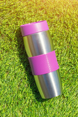 The stainless thermal flask on the green grass background with sunlight. The container for cold or hot beverages for outdoor activity. Selective focus. - 806905749
