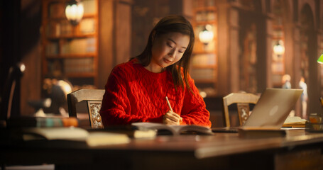 Diligent Asian Female Student Engaged in Academic Research in a Cozy Library, Writing Notes and...