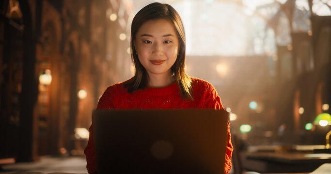 Engaged Asian Female Student Studying With Laptop in Historic Library. Young Woman Focused on E-Learning, Surrounded by Books, Preparing for College Exams in a Cozy Red Sweater.