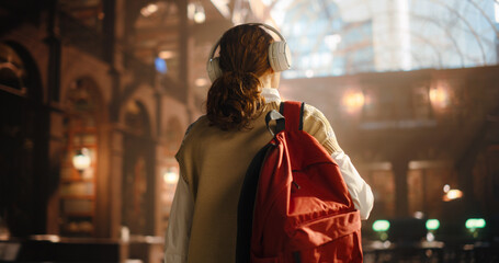 Young Female Student With Headphones and Red Backpack Walking in a Grand Library, Captured From Behind. She Is Exploring the Vast Rows of Books, Engaged in Her Academic Pursuit - Powered by Adobe