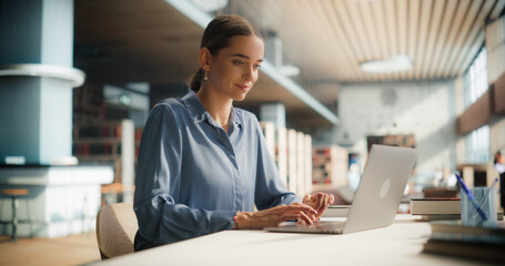 Caucasian Female Student Engaged in Academic Research on Her Laptop in a Modern Library. Young Woman Focused on Studying, Surrounded by Books and a Bright, Contemporary Educational Environment.