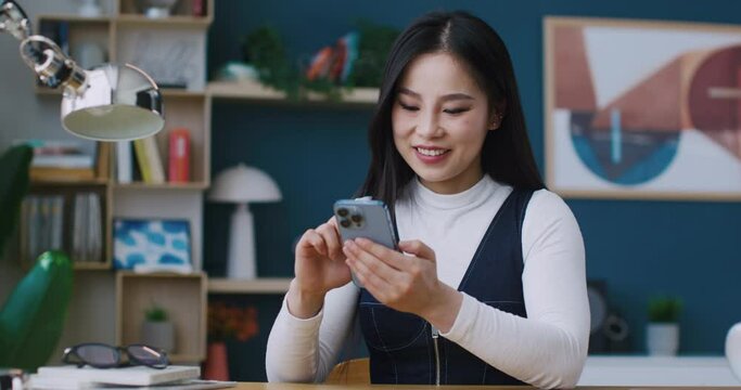 Charming Asian woman joyfully smiling while scrolling social media or texting with someone on cell phone. Exciting to spend time in Internet. Having positive feelings after watching funny video.
