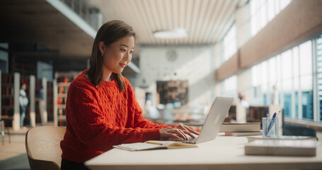 Diligent Asian Female Student Engaged in Online Learning at a Modern Library. Young Woman in Red...