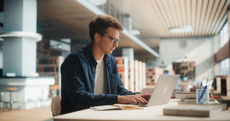 Young Man Studying in Modern Library. Caucasian Male Using Laptop Computer for College Research, Surrounded by Books and Academic Materials, Focused on Learning and Education in University Setting