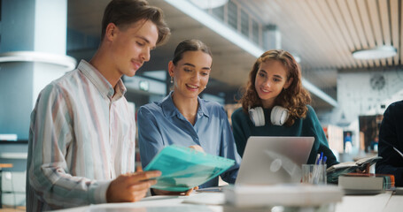 Engaged Group of University Students Collaborating on a Project in a Modern Library. Two Women and One Man Discussing Academic Material Over a Laptop and Books, Focused and Smiling.