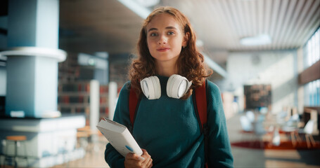 Young Caucasian Female Student With Headphones and Books Walking Through Modern Library. Confident...
