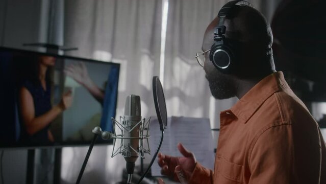 Medium side of black male voice actor in headphones reading lines from script into microphone in recording studio, while dubbing foreign movie scene of diverse couple having violent quarrel