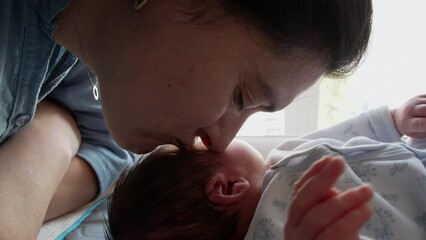 Cute maternal scene of mother kissing newborn baby by window at home during first days of life of infant son