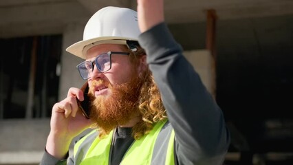 Indignant builder argues on mobile phone, gesturing with hand, discussing mistakes in planning construction work, standing against the backdrop of an unfinished building in a hard hat on a sunny day.